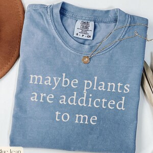May include: A blue Comfort Colors t-shirt with the phrase "maybe plants are addicted to me" in white lettering. A gold necklace with a small pendant rests on the shirt. The shirt is size small.