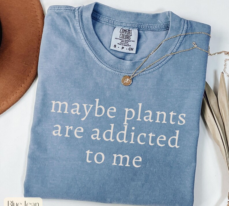 May include: A blue Comfort Colors t-shirt with the phrase "maybe plants are addicted to me" in white lettering. A gold necklace with a small pendant rests on the shirt. The shirt is size small.