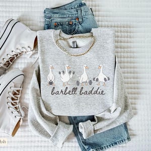 K&ouml;nnte beinhalten: Hellgraues Sweatshirt mit dem Text "barbell baddie" und Illustrationen von G&auml;nsen, die Gewichte heben. Das Sweatshirt wird mit blauen Jeans und wei&szlig;en Turnschuhen kombiniert. Eine goldene Halskette ist ebenfalls zu sehen.
