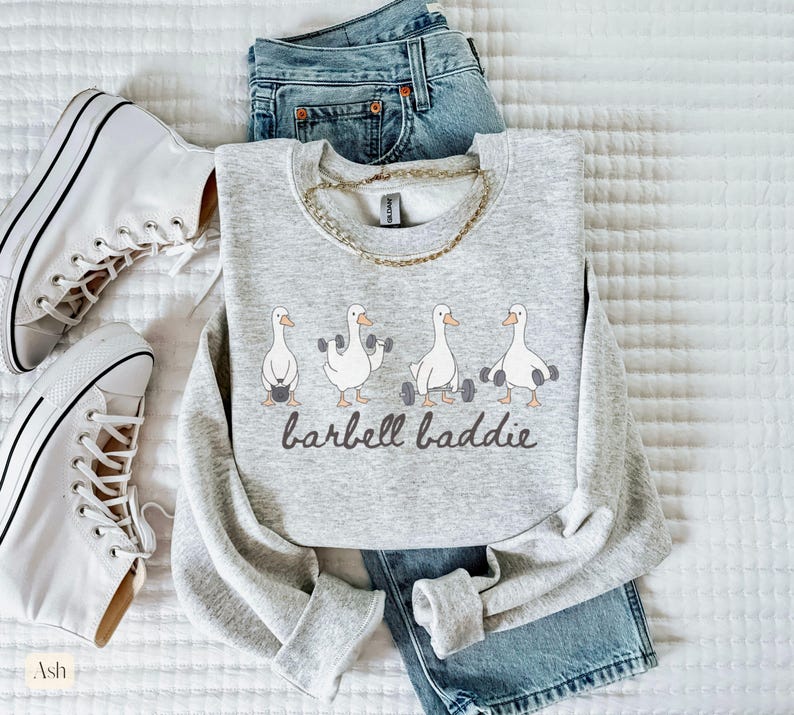 K&ouml;nnte beinhalten: Hellgraues Sweatshirt mit dem Text "barbell baddie" und Illustrationen von G&auml;nsen, die Gewichte heben. Das Sweatshirt wird mit blauen Jeans und wei&szlig;en Turnschuhen kombiniert. Eine goldene Halskette ist ebenfalls zu sehen.
