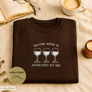 May include: Chocolate brown sweatshirt with the embroidered phrase "MAYBE WINE IS ADDICTED TO ME" and three wine glasses. A lit candle, a cup of coffee, and an open book with glasses are also in the image.