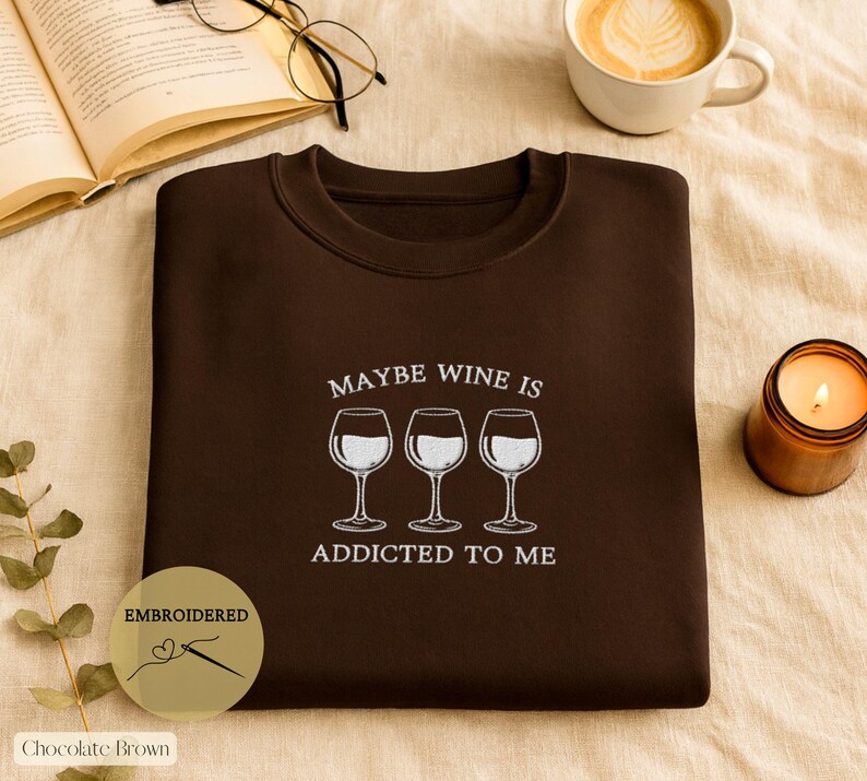 May include: Chocolate brown sweatshirt with the embroidered phrase "MAYBE WINE IS ADDICTED TO ME" and three wine glasses. A lit candle, a cup of coffee, and an open book with glasses are also in the image.