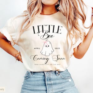 Op de afbeelding: Ivoor kleurig t-shirt met de tekst "LITTLE Boo" boven een cartoon spookje met een roze strik. Onder het spookje staan de woorden "APRIL 2025" en "Coming Soon". De tekst "BABY HUNEYCUTT ON THE WAY" staat ook op het shirt.