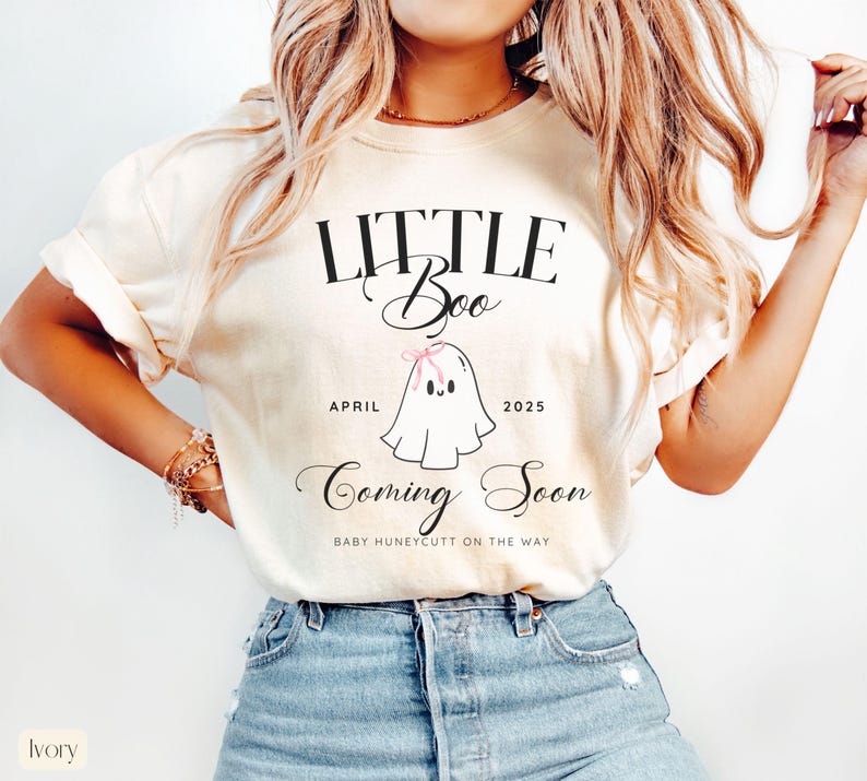 Op de afbeelding: Ivoor kleurig t-shirt met de tekst "LITTLE Boo" boven een cartoon spookje met een roze strik. Onder het spookje staan de woorden "APRIL 2025" en "Coming Soon". De tekst "BABY HUNEYCUTT ON THE WAY" staat ook op het shirt.