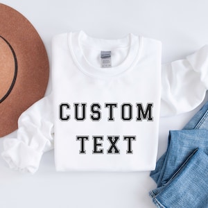 Peut inclure: Sweat-shirt blanc avec un texte noir qui dit "CUSTOM TEXT" dans une police de caract&egrave;res de style universitaire.
