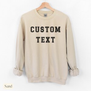 Peut inclure: Un sweat-shirt couleur sable avec un texte noir qui dit "CUSTOM TEXT" dans une police de caract&egrave;res de style universitaire.
