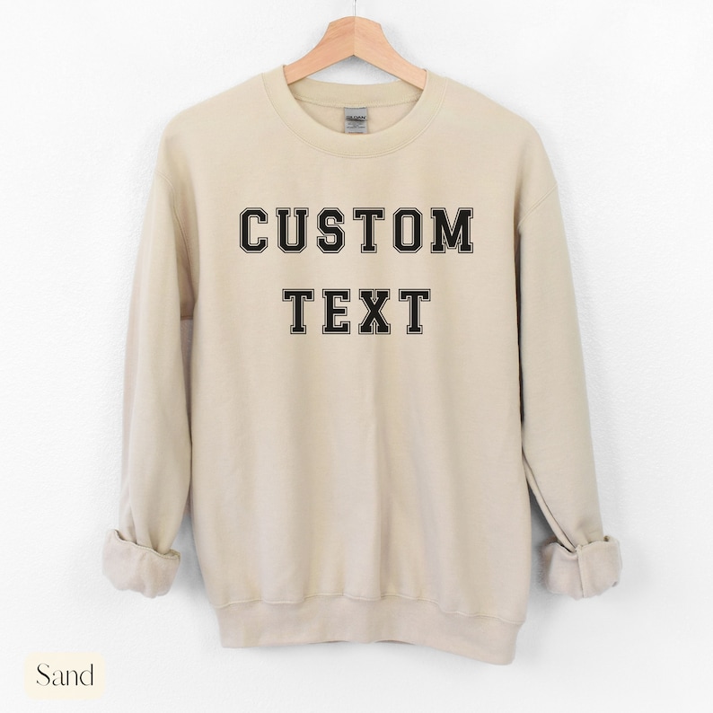 Peut inclure: Un sweat-shirt couleur sable avec un texte noir qui dit "CUSTOM TEXT" dans une police de caract&egrave;res de style universitaire.