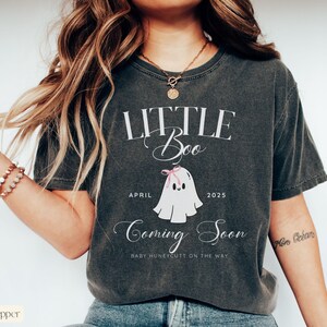 Op de afbeelding: Donkergrijs t-shirt met de tekst "LITTLE Boo" en "Coming Soon". Een cartoon spookje met een roze strik staat onder de tekst, samen met de maand "April" en het jaar "2025". De zin "BABY HUNEYCUTT ON THE WAY" is ook opgenomen.