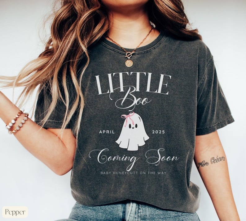 Op de afbeelding: Donkergrijs t-shirt met de tekst "LITTLE Boo" en "Coming Soon". Een cartoon spookje met een roze strik staat onder de tekst, samen met de maand "April" en het jaar "2025". De zin "BABY HUNEYCUTT ON THE WAY" is ook opgenomen.