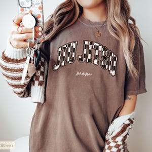 Pu&ograve; includere: T-shirt color espresso con le parole "JIU JITSU" in un motivo a scacchi, arcuate sul petto. Sotto il testo c'&egrave; la parola "mom" in un carattere pi&ugrave; piccolo. La maglietta &egrave; di vestibilit&agrave; ampia.