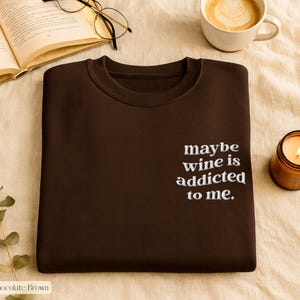 Puede incluir: Sudadera marr&oacute;n chocolate con la frase bordada "maybe wine is addicted to me." La imagen tambi&eacute;n incluye una vela encendida, una taza de caf&eacute; y un libro abierto con gafas.