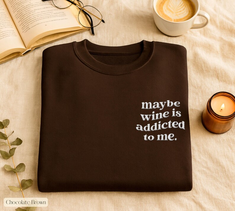 Puede incluir: Sudadera marr&oacute;n chocolate con la frase bordada "maybe wine is addicted to me." La imagen tambi&eacute;n incluye una vela encendida, una taza de caf&eacute; y un libro abierto con gafas.