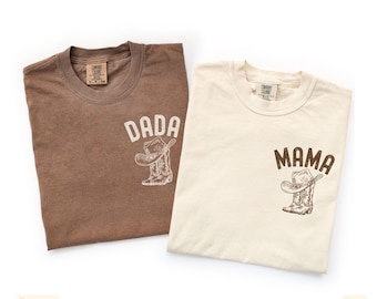 Puede incluir: Se muestran dos camisetas, una espresso y otra marfil. La camiseta espresso tiene "DADA" impreso sobre un gráfico de bota y sombrero de vaquero. La camiseta marfil tiene "MAMA" impresa sobre un gráfico similar. Ambas son de manga corta.
