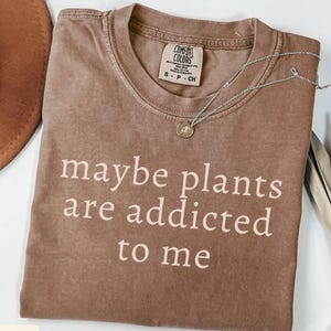 May include: A brown Comfort Colors t-shirt with the text "maybe plants are addicted to me" in white. A silver necklace with a small pendant is draped across the shirt. A brown hat is visible in the upper left corner.