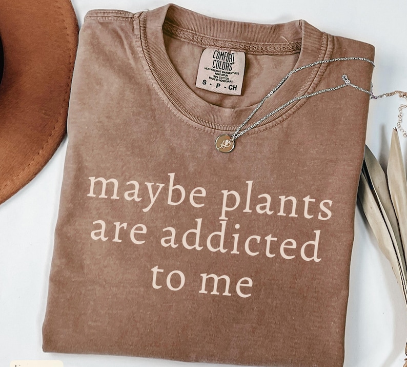 May include: A brown Comfort Colors t-shirt with the text "maybe plants are addicted to me" in white. A silver necklace with a small pendant is draped across the shirt. A brown hat is visible in the upper left corner.