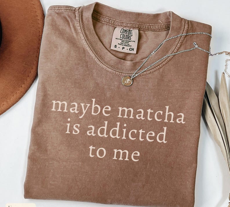 Puede incluir: Una camiseta marr&oacute;n Comfort Colors con el texto "maybe matcha is addicted to me" en blanco. Un collar plateado con un peque&ntilde;o colgante est&aacute; sobre la camiseta. La camiseta es talla small.