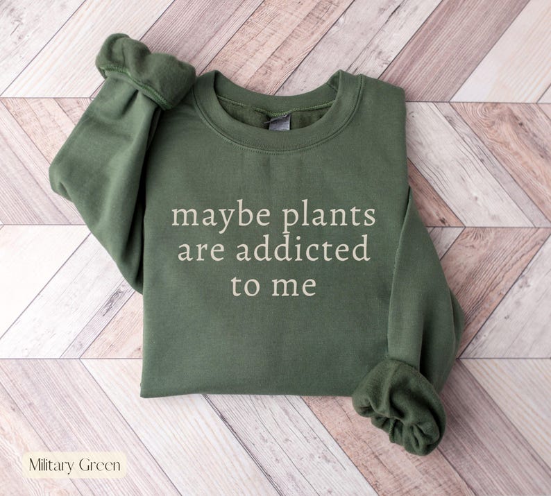 May include: Military green sweatshirt with the text "maybe plants are addicted to me" in white. The long-sleeved crew neck top is made of a soft material. The sweatshirt is a casual style.