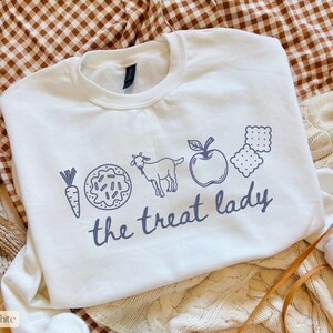 Puede incluir: Sudadera blanca con cuello redondo con un gr&aacute;fico azul de una zanahoria, una rosquilla, una cabra, una manzana y galletas saladas. Las palabras "the treat lady" est&aacute;n impresas debajo.