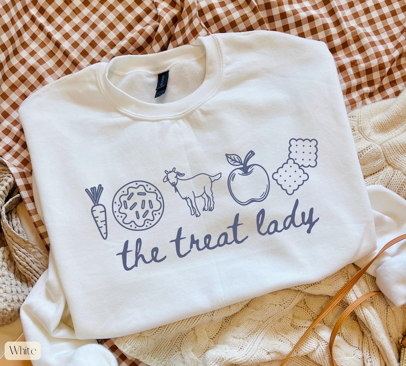 Puede incluir: Sudadera blanca con cuello redondo con un gr&aacute;fico azul de una zanahoria, una rosquilla, una cabra, una manzana y galletas saladas. Las palabras "the treat lady" est&aacute;n impresas debajo.
