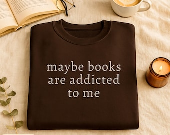 Puede incluir: Sudadera marrón chocolate con el texto blanco "maybe books are addicted to me". La imagen también incluye un libro abierto, gafas, una taza de café y una vela encendida.