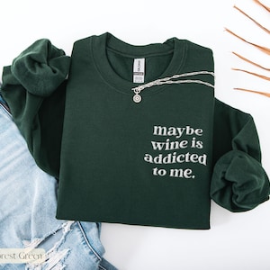Puede incluir: Sudadera verde bosque con la frase bordada "maybe wine is addicted to me." Un collar plateado con un colgante est&aacute; colocado sobre la sudadera. La sudadera est&aacute; junto a unos vaqueros azules.