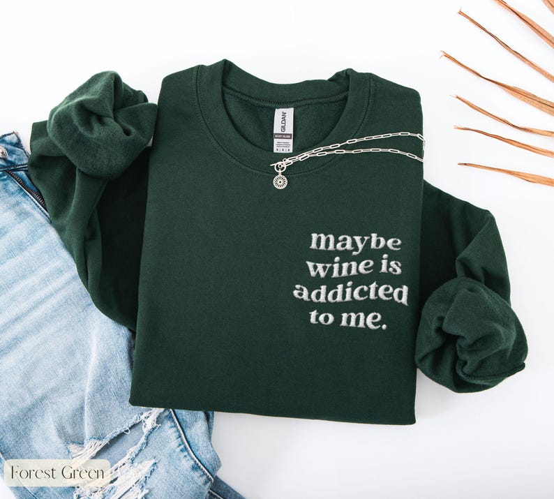 Puede incluir: Sudadera verde bosque con la frase bordada "maybe wine is addicted to me." Un collar plateado con un colgante est&aacute; colocado sobre la sudadera. La sudadera est&aacute; junto a unos vaqueros azules.