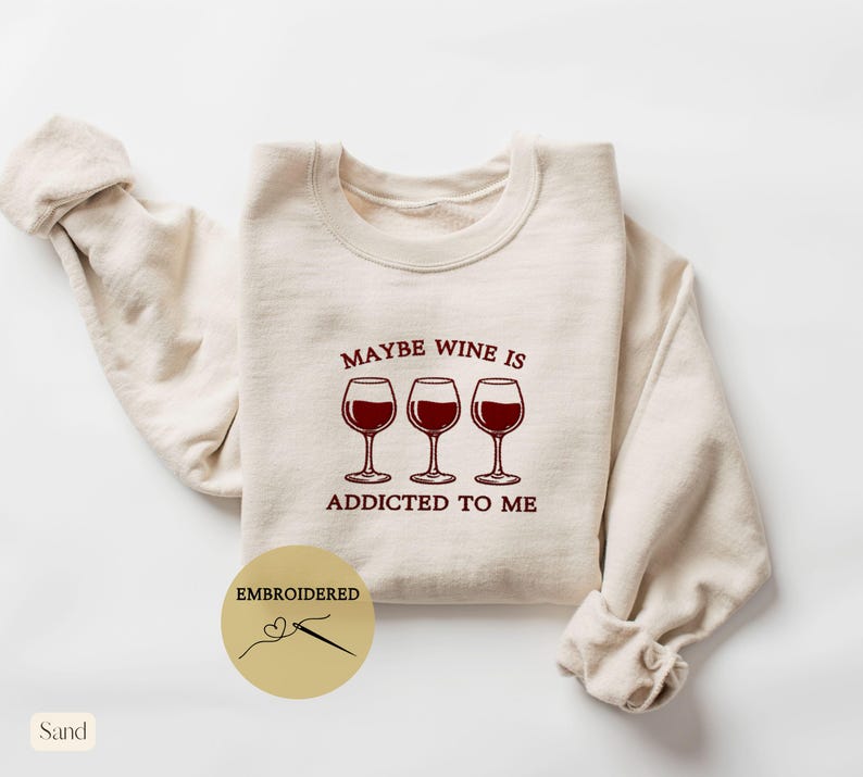 May include: Sand-colored sweatshirt with the embroidered phrase "MAYBE WINE IS ADDICTED TO ME" above three wine glasses filled with red wine. The word "EMBROIDERED" is printed on a gold circle with a needle and thread design.