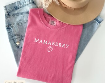 May include: A pink t-shirt with the word "MAMABERRY" and a strawberry graphic printed in white. The shirt is folded on top of a pair of blue jeans, with a tan hat and a gold necklace visible.