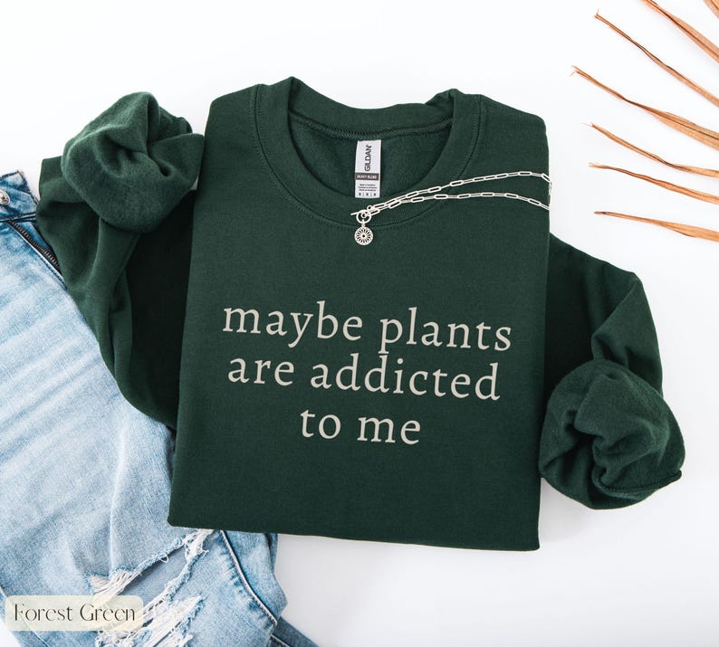 May include: Forest green sweatshirt with the text "maybe plants are addicted to me" in white. A silver necklace with a pendant is draped across the sweatshirt. The sweatshirt is folded next to a pair of blue jeans.