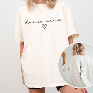 K&ouml;nnte beinhalten: &Uuml;bergr&ouml;&szlig;en-T-Shirt in Elfenbein mit dem Text "dance mama" und einem Herz auf der Vorderseite. Die R&uuml;ckseite des Shirts tr&auml;gt den Text "Theodore" mit einem Herz auf jeder Seite.
