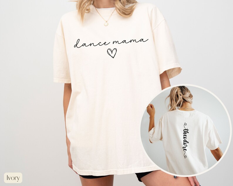 K&ouml;nnte beinhalten: &Uuml;bergr&ouml;&szlig;en-T-Shirt in Elfenbein mit dem Text "dance mama" und einem Herz auf der Vorderseite. Die R&uuml;ckseite des Shirts tr&auml;gt den Text "Theodore" mit einem Herz auf jeder Seite.