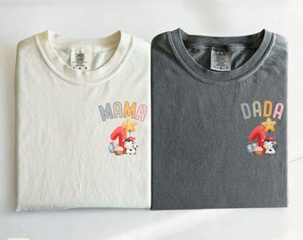 Puede incluir: Dos camisetas dobladas, una marfil y otra pimienta, con las palabras "MAMA" y "DADA" impresas. Cada camiseta tiene un gráfico colorido con el número uno, una estrella y una vaca. La camiseta marfil está etiquetada como "Ivory" y la camiseta pimienta como "Pepper".