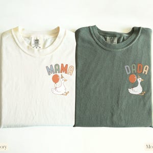 Puede incluir: Dos camisetas Comfort Colors, marfil y verde musgo, con gr&aacute;ficos a juego. Cada camiseta presenta un ganso de dibujos animados sosteniendo un globo, con las palabras "MAMA" y "DADA" encima del ganso. La camiseta marfil est&aacute; etiquetada como "Ivory" y la camiseta verde como "Moss".