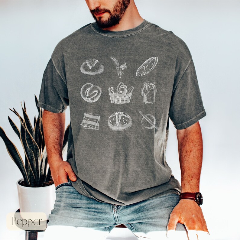 Peut inclure: Un t-shirt gris avec un motif vintage repr&eacute;sentant diff&eacute;rents types de pain, un panier et un bocal. Le motif est en blanc et est centr&eacute; sur le t-shirt.
