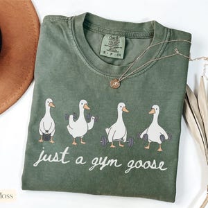 K&ouml;nnte beinhalten: Moosgr&uuml;nes T-Shirt mit Rundhalsausschnitt und einem Aufdruck von vier wei&szlig;en Cartoon-G&auml;nsen, die Gewichte heben. Der Text "just a gym goose" ist darunter in Schreibschrift gedruckt. Eine goldene Halskette liegt auf dem Shirt.