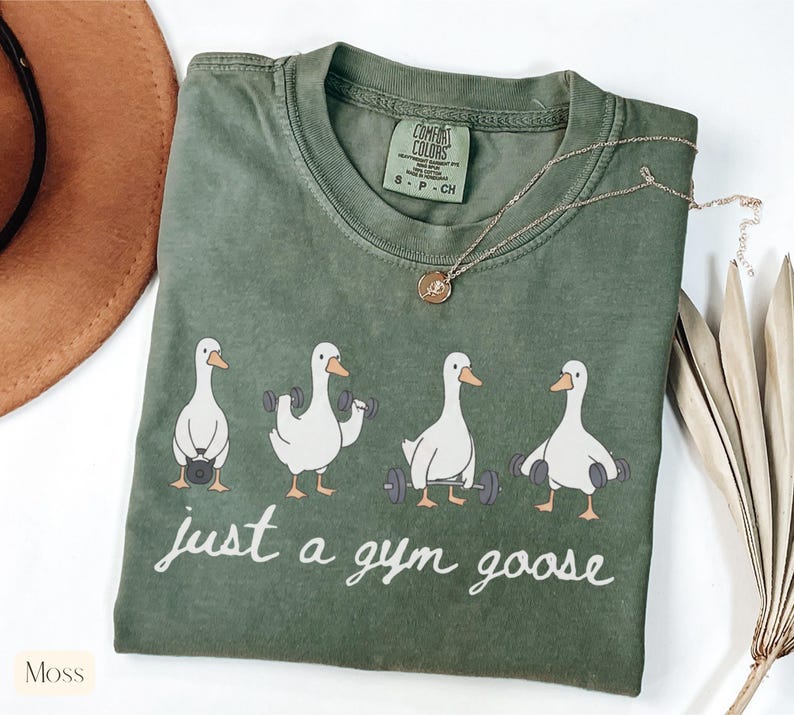 K&ouml;nnte beinhalten: Moosgr&uuml;nes T-Shirt mit Rundhalsausschnitt und einem Aufdruck von vier wei&szlig;en Cartoon-G&auml;nsen, die Gewichte heben. Der Text "just a gym goose" ist darunter in Schreibschrift gedruckt. Eine goldene Halskette liegt auf dem Shirt.