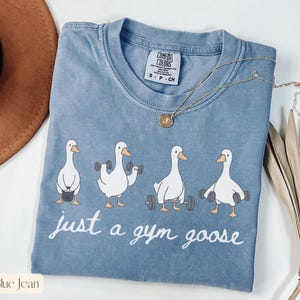 K&ouml;nnte beinhalten: Blaues Jeans-T-Shirt mit einer Grafik von vier Cartoon-G&auml;nsen, die Gewichte heben. Der Text "just a gym goose" steht in wei&szlig;er Schrift darunter. Eine goldene Halskette liegt auf dem T-Shirt.