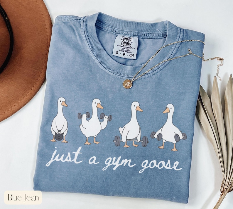 K&ouml;nnte beinhalten: Blaues Jeans-T-Shirt mit einer Grafik von vier Cartoon-G&auml;nsen, die Gewichte heben. Der Text "just a gym goose" steht in wei&szlig;er Schrift darunter. Eine goldene Halskette liegt auf dem T-Shirt.