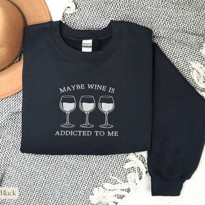 May include: A black sweatshirt with the text "MAYBE WINE IS ADDICTED TO ME" and three wine glasses in white. The sweatshirt is on a patterned blanket with a brown hat in the background.