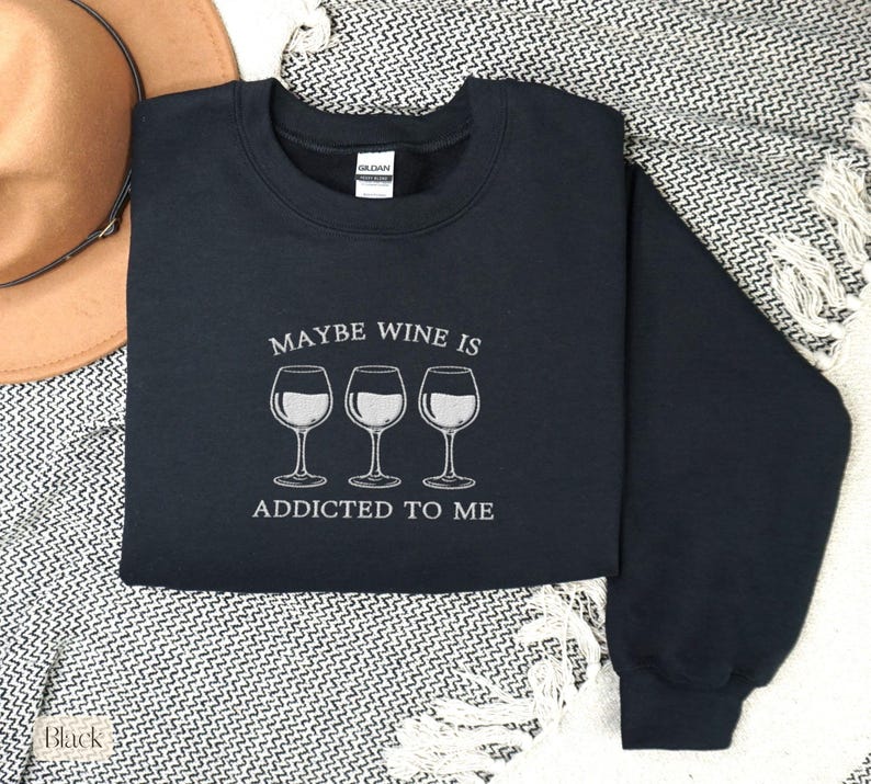 May include: A black sweatshirt with the text "MAYBE WINE IS ADDICTED TO ME" and three wine glasses in white. The sweatshirt is on a patterned blanket with a brown hat in the background.