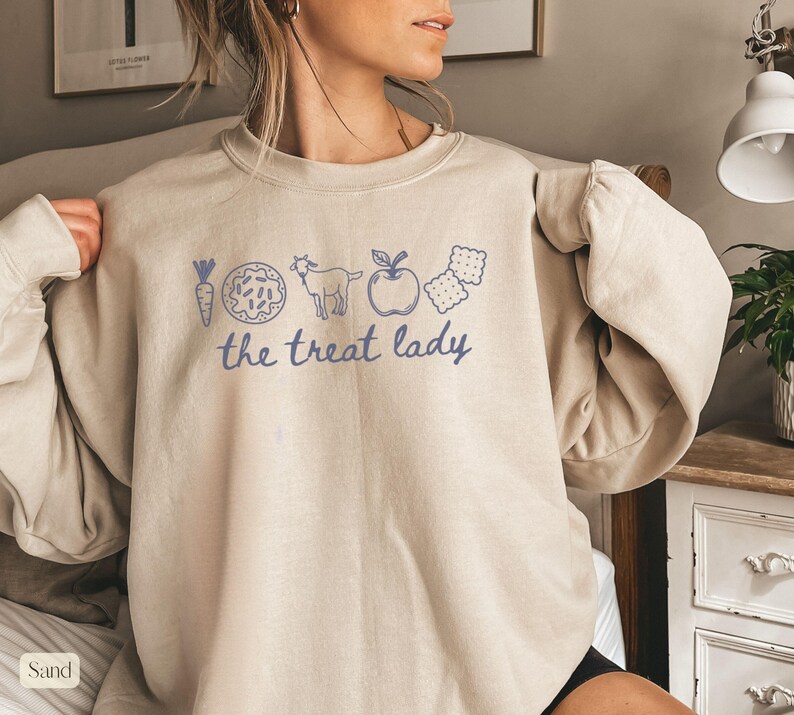 Puede incluir: Sudadera color arena con un gr&aacute;fico azul de una zanahoria, una rosquilla, una cabra, una manzana y galletas, con el texto "the treat lady". La sudadera tiene mangas largas y cuello redondo.
