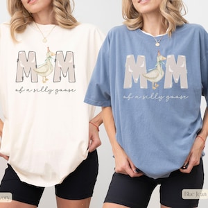 Puede incluir: Dos camisetas con el texto "MOM of a silly goose" y un ganso de dibujos animados con un gorro de fiesta. La camiseta de la izquierda es de color marfil y la camiseta de la derecha es de color azul vaquero.