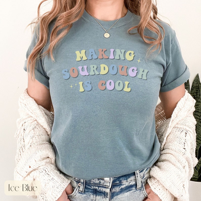 Peut inclure: Un t-shirt bleu clair avec le texte "Making sourdough is cool" en lettres color&eacute;es de style r&eacute;tro.