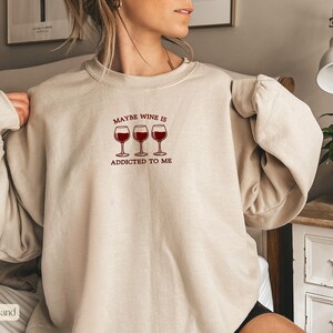 May include: Sand-colored sweatshirt with the text "Maybe Wine Is Addicted To Me" above three wine glasses filled with red wine. The sweatshirt has long sleeves and a crew neck.