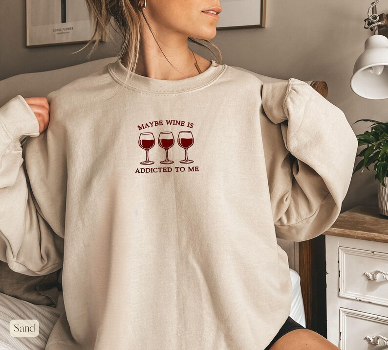 May include: Sand-colored sweatshirt with the text "Maybe Wine Is Addicted To Me" above three wine glasses filled with red wine. The sweatshirt has long sleeves and a crew neck.