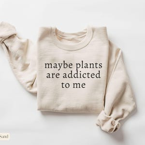 May include: Sand-colored sweatshirt with the text "maybe plants are addicted to me" in black. The long-sleeved top is folded, with the sleeves rolled up. The word "Sand" is in a small rectangle in the bottom left corner.