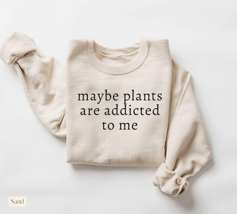 May include: Sand-colored sweatshirt with the text "maybe plants are addicted to me" in black. The long-sleeved top is folded, with the sleeves rolled up. The word "Sand" is in a small rectangle in the bottom left corner.