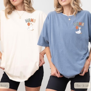 Puede incluir: Camisetas color marfil y azul vaquero con la palabra "MAMA" y un dibujo de un ganso con un globo. La camiseta marfil tiene pantalones cortos negros, y la camiseta azul vaquero tiene pantalones cortos negros. Las camisetas son de manga corta y de corte holgado.