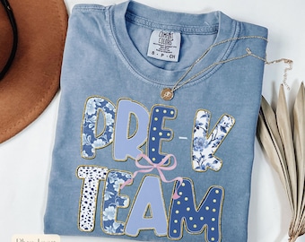 Puede incluir: Una camiseta azul vaquera con las palabras "PRE-K TEAM" en una mezcla de letras florales azules, lunares y azules lisas. Un lazo rosa está entre la "E" y la "T". Un collar dorado está en la camisa.