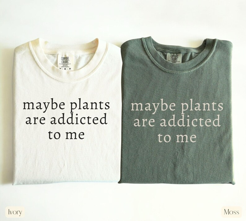 May include: Two t-shirts, one ivory and one moss green, are displayed. Each shirt has the phrase "maybe plants are addicted to me" printed in a contrasting color. The ivory shirt has black text, while the moss shirt has white text.